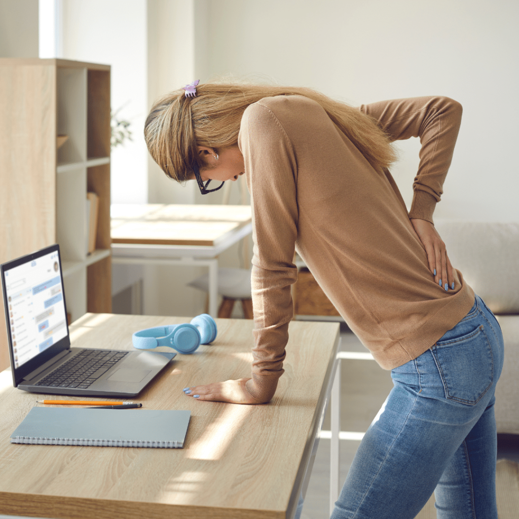 Woman supporting herself by leaning on her desk while holding her lower back in pain.