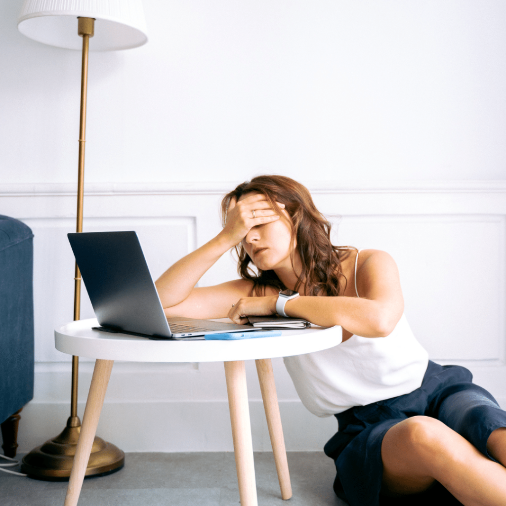 Lady sitting on ground leaned onto coffee table with hands covering eyes and forehead, laptop sitting on desk.