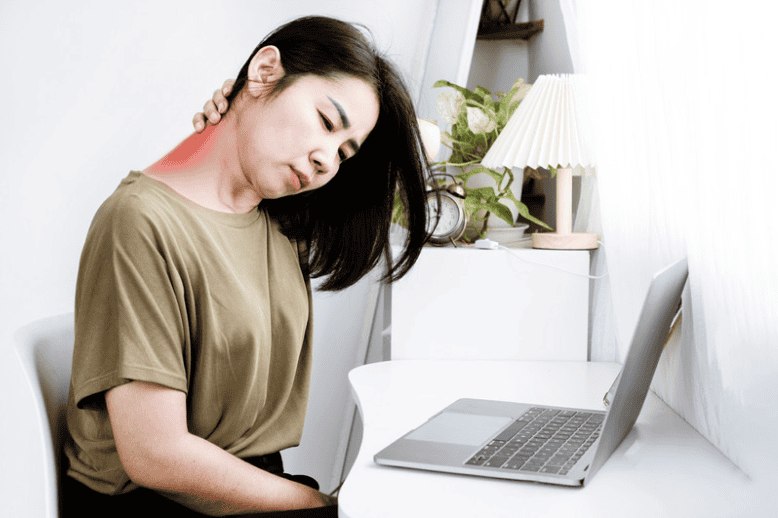 Lady sitting at her desk leaning head over while holding hand onto back of neck, sitting in front of a laptop inside her room.