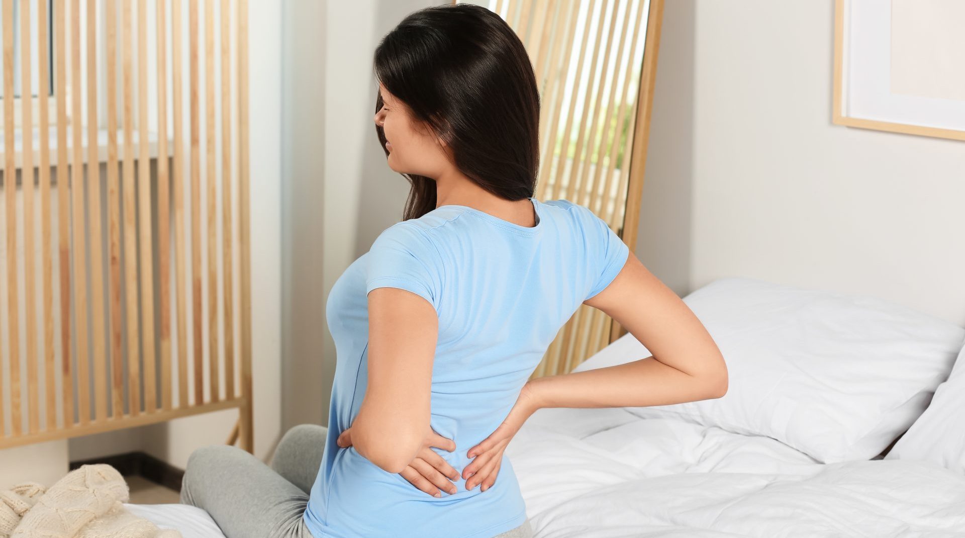Woman sitting on a bed holding her lower back with both hands, indicating back pain or discomfort after waking up.