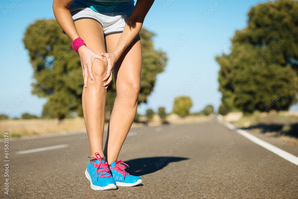 Female runner holding her knee in pain on a rural road, suggesting knee injury or joint discomfort during exercise.