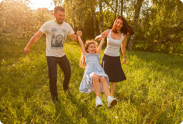 Happy family enjoying time outdoors, parents swinging their smiling daughter in a sunny green park.