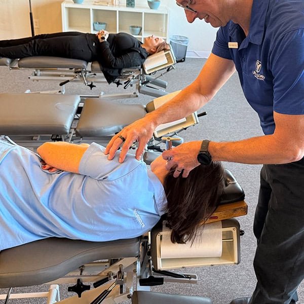 Chiropractor adjusting a female patient's neck in a side-lying position while another patient relaxes in the background on a treatment table.