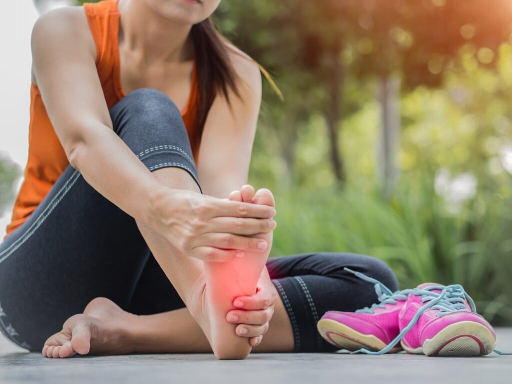 Female runner sitting outdoors holding her foot in pain, highlighting plantar fasciitis or heel injury.