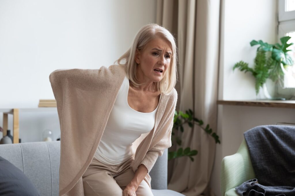 Older woman with blonde hair holding her lower back in pain while sitting on a couch at home, showing signs of discomfort or a possible back injury.