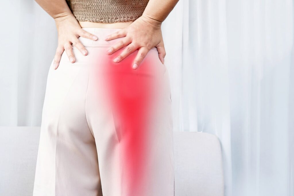 Woman holding her lower back with red highlighted pain radiating down her leg, indicating sciatica or nerve pain.