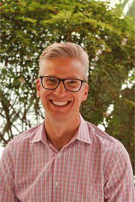 Dr. Cox, founder and chiropractor, smiling outdoors in a pink checkered shirt with greenery in the background.