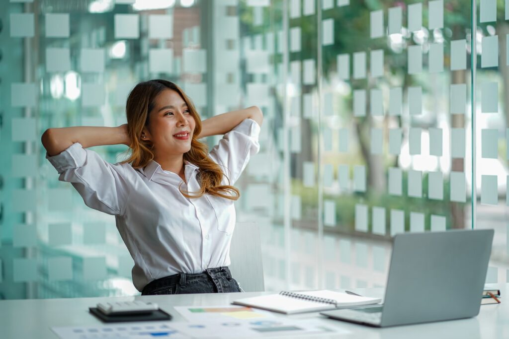 A relaxed young woman in a white shirt leans back with her hands behind her head, smiling while sitting at a desk with a laptop and documents in a modern glass-walled office.