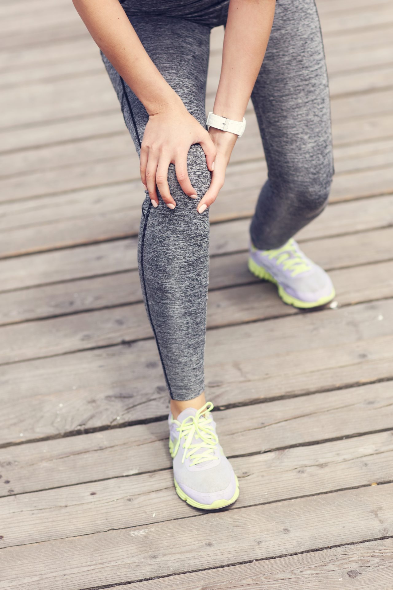 Woman in athletic clothing holding her knee in discomfort while standing outdoors.