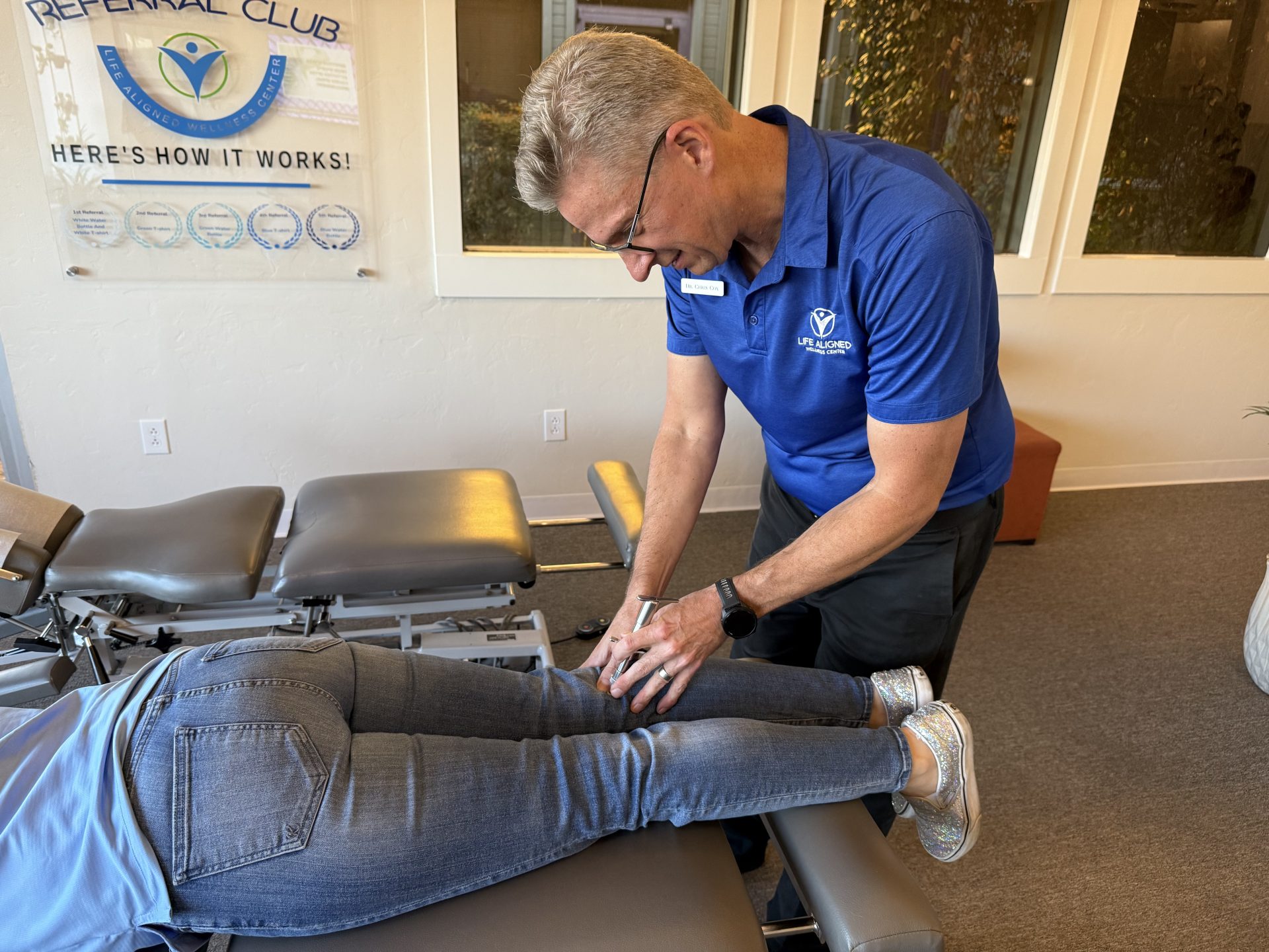 Chiropractor applying an adjusting instrument to a patient’s lower leg while she lies face down.